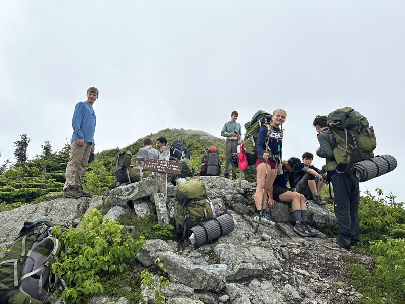 A group of people are gathered on a rocky mountaintop, likely after a hike. They are surrounded by backpacks and gear, suggesting they have been trekking. The sky is overcast, creating a somewhat hazy atmosphere. The group appears to be resting and enjoying the view from their elevated vantage point.